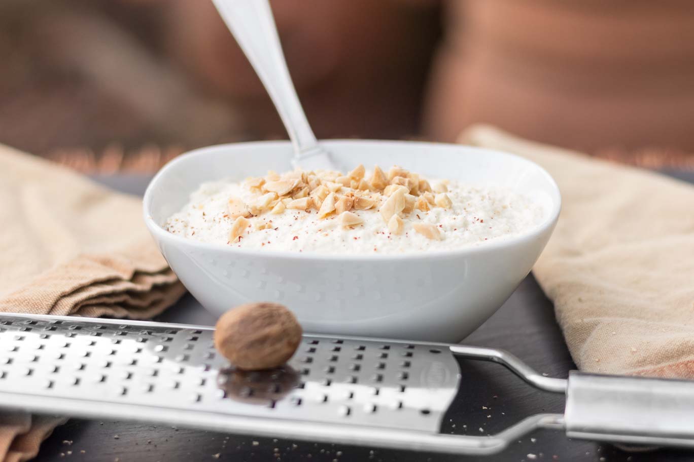 A micro plane grater with a nutmeg ball on top and the Creamy Almond Ricotta in a small bowl garnished with toasted almonds, nutmeg and fresh ground pepper behind.