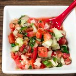 Small square white bowl Garden Salsa Fresca is a naturally sos free salsa fresca with fresh tomatoes, white onion, cucumbers, cilantro, burst of lime juice and a little kick from the jalapeno pepper. with a red spoon.