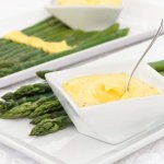 Silky smooth oil-free vegan hollandaise in a square white bowl served with blanched asparagus on white rectangular plates.