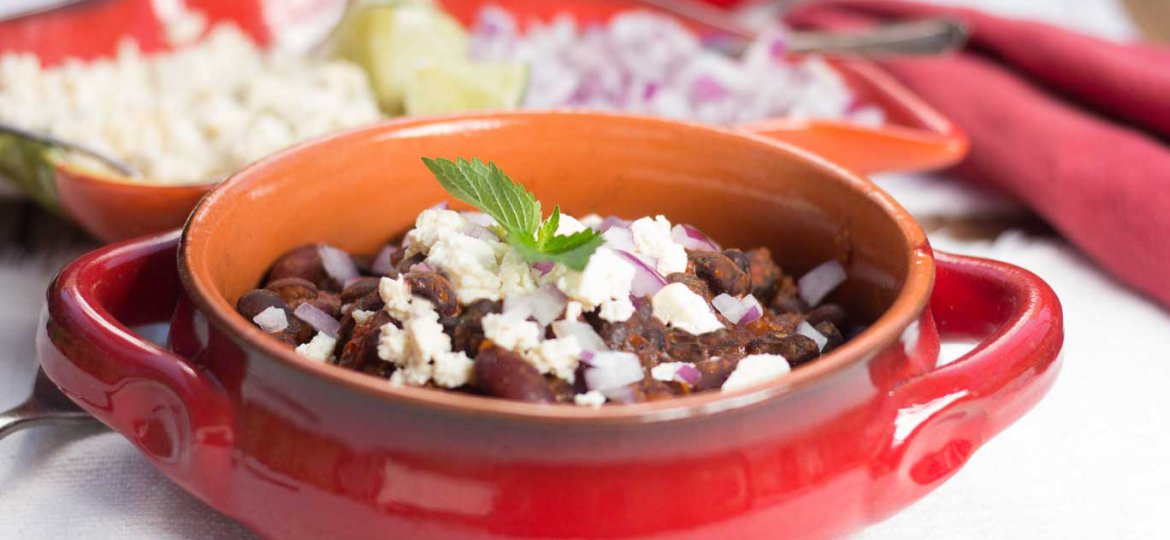 Red-glazed terracotta casserole serving dish with Chile Bean ChiliOil-free Vegan Chile Bean Chili and a chile pepper-shaped dish with tangy bites, lime wedges and diced red onion behind.