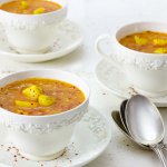 Three white wedgewood cups of Red Lentil Soup with Carrot and Ginger garnished with spice potatoes and a sprinkle of Aleppo pepper.