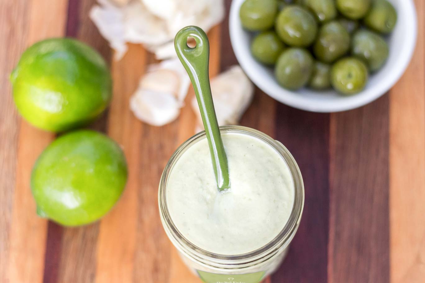 An open jar of “Olivaise” Oil-free Vegan Mayonnaise surrounded by limes, garlic cloves and green olives.