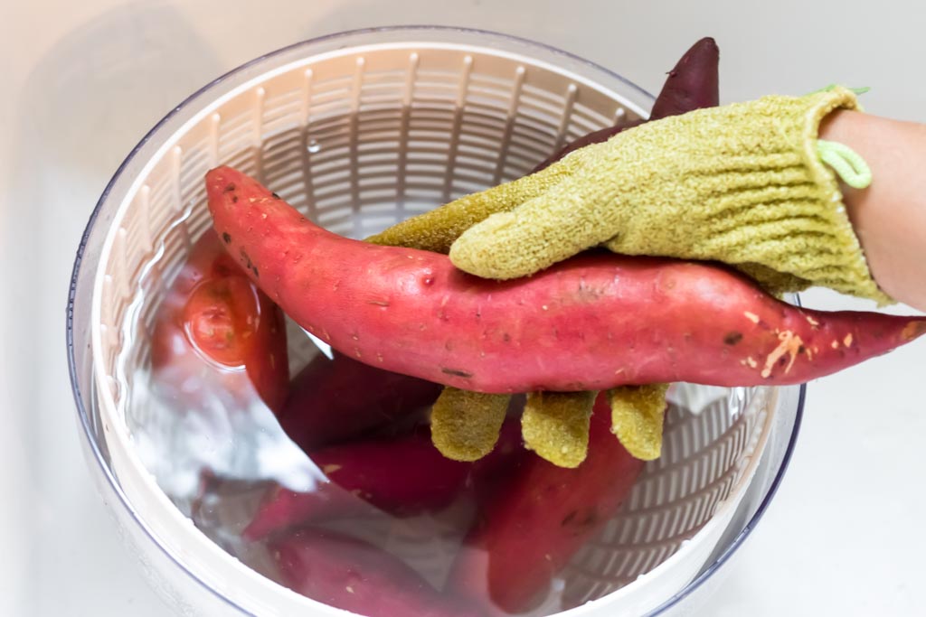 Hand with scrub glove holding a sweet potato above the salad spinner with water