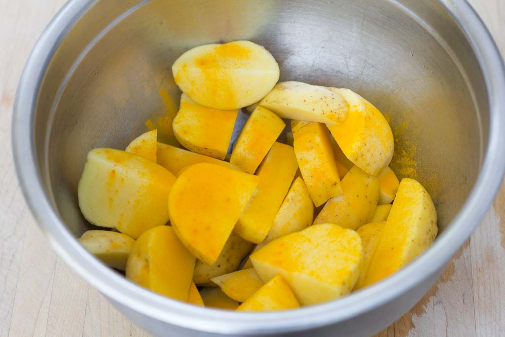 Stainless steel prep bowl with cut potatoes and first sprinkle of turmeric powder