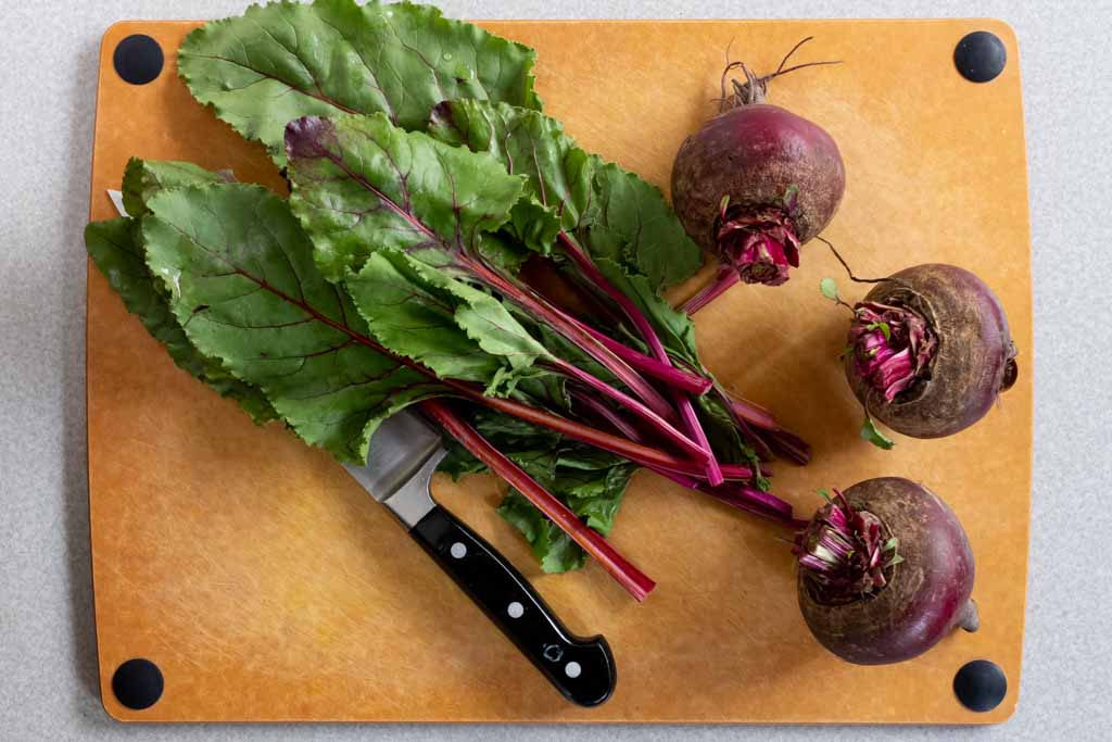 Three Dark Red Beets with their greens just cut on a cutting board with a chef knife