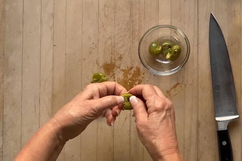 Removing pit from the Castelvetrano olives on a large cutting board with a small glass bowl to hold the pitted olives