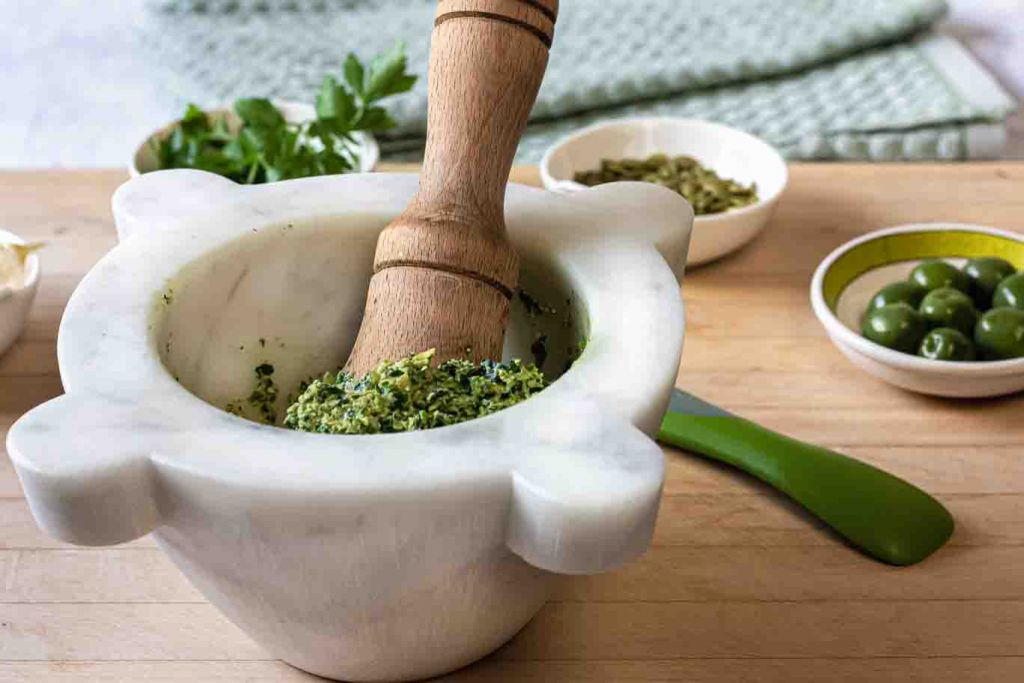 Oil-free Vegan Parsley pesto in the marble mortar with the wood pestle on a cutting board small bowls of Castelvetrano olives, pumpkin seeds and parsley leaves